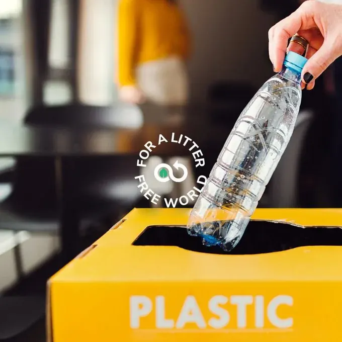 Person placing a plastic bottle into a yellow plastic recycling bin labeled plastic